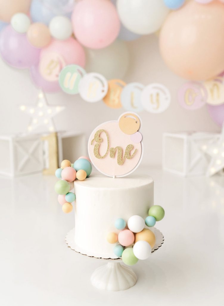 Bubbly One first birthday cake topper with pastel balloon garland on white cake stand
